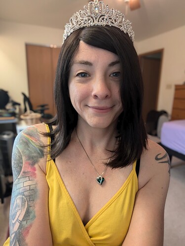 A person with a tiara on their head is smiling at the camera, wearing a yellow sleeveless top, and has visible tattoos on their arms. (Captioned by AI)